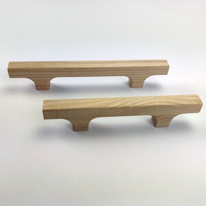 Puede incluir: Dos tiradores de madera sin terminar. Cada tirador es rectangular con un asa curvada y dos peque&ntilde;os pies. La veta de la madera es visible y los tiradores son de color marr&oacute;n claro. Adecuado para la restauraci&oacute;n de muebles o proyectos de bricolaje.