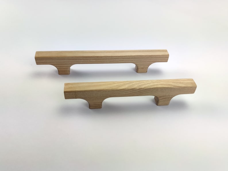 Puede incluir: Dos tiradores de madera sin terminar. Cada tirador es rectangular con un asa curvada y dos peque&ntilde;os pies. La veta de la madera es visible y los tiradores son de color marr&oacute;n claro. Adecuado para la restauraci&oacute;n de muebles o proyectos de bricolaje.