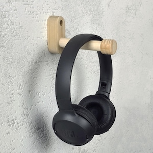 May include: A wooden headphone holder fixed to a textured grey wall. The holder is made of light-coloured wood and supports a pair of black headphones. The headphones have the brand name "JBL" printed on them.
