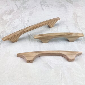 May include: Three wooden cabinet handles in varying sizes. The handles are light brown with a natural wood grain. The design features a curved shape with a flat top and a slightly flared base. The handles are designed for easy grip and installation.