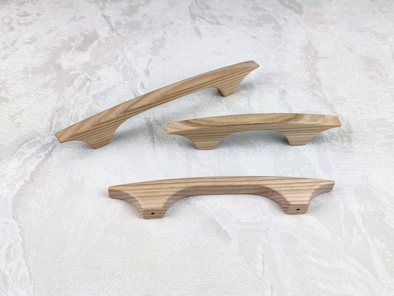 May include: Three wooden cabinet handles in varying sizes. The handles are light brown with a natural wood grain. The design features a curved shape with a flat top and a slightly flared base. The handles are designed for easy grip and installation.