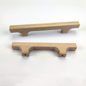 Puede incluir: Dos tiradores de armario de madera clara. Cada tirador es rectangular con un asa curvada y dos agujeros para el montaje. La veta de la madera es visible y los tiradores est&aacute;n sobre un fondo blanco.