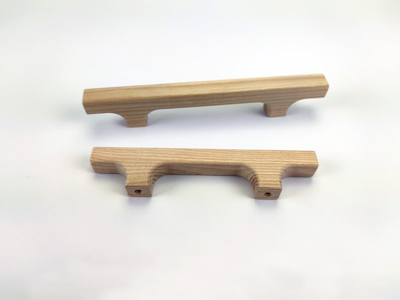 Puede incluir: Dos tiradores de armario de madera clara. Cada tirador es rectangular con un asa curvada y dos agujeros para el montaje. La veta de la madera es visible y los tiradores est&aacute;n sobre un fondo blanco.