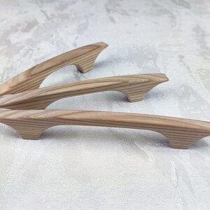 May include: Three wooden cabinet pulls with a natural wood grain finish. The pulls have a curved handle design with flared ends. The wood has a light brown colour. The pulls are approximately 15 cm long.