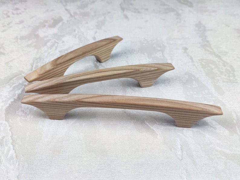 May include: Three wooden cabinet pulls with a natural wood grain finish. The pulls have a curved handle design with flared ends. The wood has a light brown colour. The pulls are approximately 15 cm long.