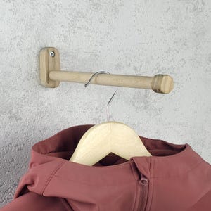 May include: A wooden coat rack with a cylindrical bar and a small wooden mounting plate. A silver metal hook is attached to the bar. A wooden hanger holds a maroon jacket. The rack is mounted on a textured grey wall.