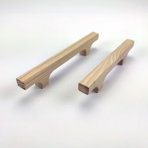 Puede incluir: Dos tiradores de madera para armarios con acabado natural. Los tiradores son rectangulares con un agarre curvo y dos peque&ntilde;os pies. La veta de la madera es visible. Un tirador es ligeramente m&aacute;s grande que el otro.
