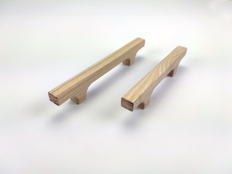 Puede incluir: Dos tiradores de madera para armarios con acabado natural. Los tiradores son rectangulares con un agarre curvo y dos peque&ntilde;os pies. La veta de la madera es visible. Un tirador es ligeramente m&aacute;s grande que el otro.