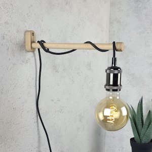 Op de afbeelding: Een houten wandlamp met een zwart snoer en een grote, ronde gloeilamp in Edison-stijl. De armatuur is gemaakt van lichtgekleurd hout en heeft een eenvoudig, minimalistisch ontwerp.