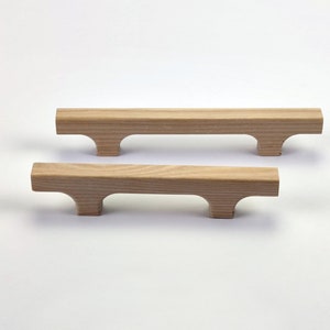 Puede incluir: Dos tiradores de armario de madera con acabado natural. Cada tirador tiene forma de barra rectangular con un recorte curvo en cada extremo, creando un asa. Los tiradores son de madera de color claro y est&aacute;n sobre un fondo blanco.