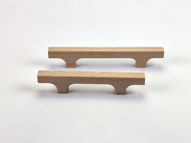 Puede incluir: Dos tiradores de armario de madera con acabado natural. Cada tirador tiene forma de barra rectangular con un recorte curvo en cada extremo, creando un asa. Los tiradores son de madera de color claro y est&aacute;n sobre un fondo blanco.