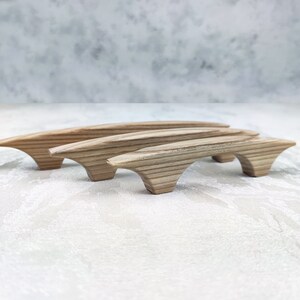 May include: Three wooden cabinet pulls with a natural wood grain finish. The pulls have a curved design with a bridge-like shape, and are arranged in a row. The pulls are made of light-coloured wood.