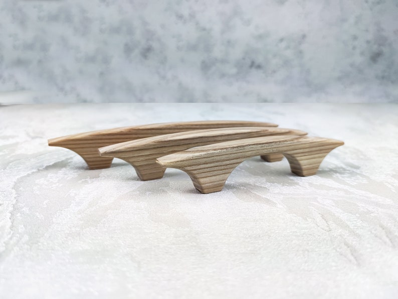 May include: Three wooden cabinet pulls with a natural wood grain finish. The pulls have a curved design with a bridge-like shape, and are arranged in a row. The pulls are made of light-coloured wood.