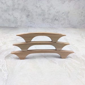 May include: Three light-coloured wooden cabinet pulls are stacked. Each pull has a curved, bridge-like design with flared ends. The wood grain is visible, and the pulls are set against a neutral background. These pulls are approximately 15 cm long.