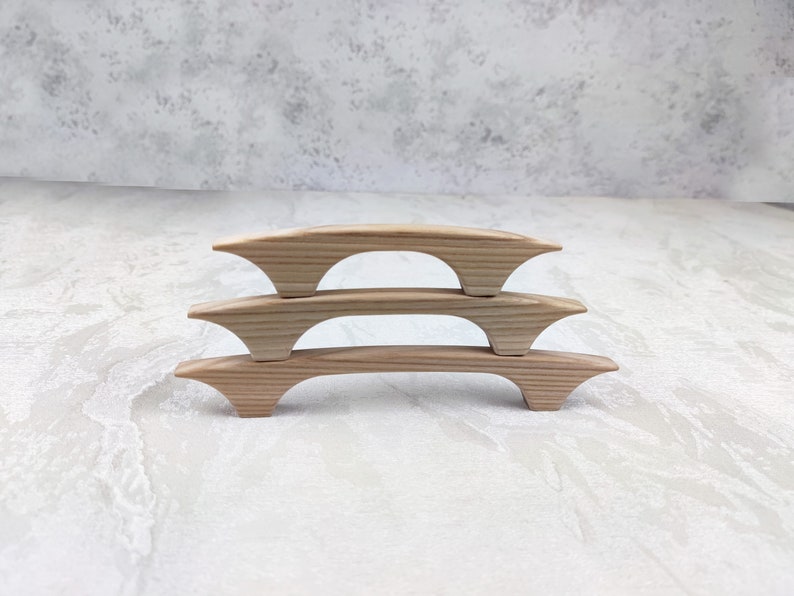 May include: Three light-coloured wooden cabinet pulls are stacked. Each pull has a curved, bridge-like design with flared ends. The wood grain is visible, and the pulls are set against a neutral background. These pulls are approximately 15 cm long.