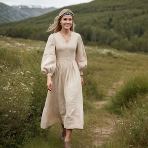 Puede incluir: Un vestido de lino beige con cuello en V y mangas largas abullonadas. El vestido tiene una cintura ajustada y una falda fluida. La mujer lleva una diadema estampada y camina por un sendero en un campo.