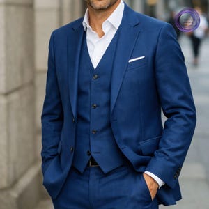 May include: A three-piece, navy blue suit with a white dress shirt. The suit includes a blazer, vest, and trousers. The blazer has a pocket square and the vest has buttons. The person is standing outside.