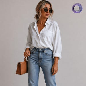 May include: A woman wearing a white button-down shirt, light blue jeans, and sunglasses. She is carrying a brown handbag. The shirt has a pocket on the chest. The outfit is casual and stylish.