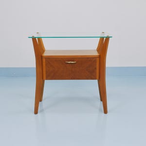 May include: A mid-century modern nightstand with a glass top and a single drawer. The drawer and frame are made of light-colored wood with a decorative pattern on the drawer front. The legs are slender and angled.
