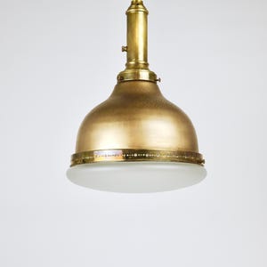 May include: A vintage-style brass pendant light fixture. The light has a domed shade with a frosted glass diffuser. The fixture is suspended from a brass rod and ceiling mount. The overall design is a classic, elegant style.