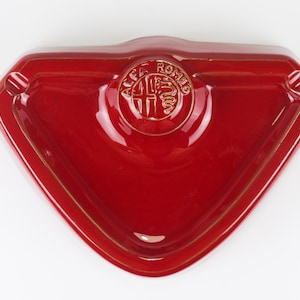 May include: A glossy red ceramic ashtray shaped like a triangular shield with rounded corners. The center features a raised circular emblem with the embossed text ALFA ROMEO, depicting a cross on the left and a serpent on the right side.