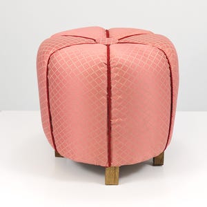 May include: A square, upholstered ottoman in a soft coral color with a diamond pattern. The seams are accented with a darker red trim. The ottoman sits on four short, square wooden legs.