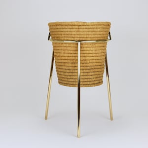 May include: A woven basket planter with a cylindrical shape, supported by a gold-colored metal stand with three legs. The basket is made of a light brown, textured material. The stand is a sleek, minimalist design.