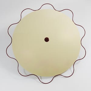 May include: A round, cream-colored ceiling light fixture with a wavy, dark red metal frame. The light has a small, dark red center detail. The fixture is designed to be mounted on a ceiling and provides ambient lighting.