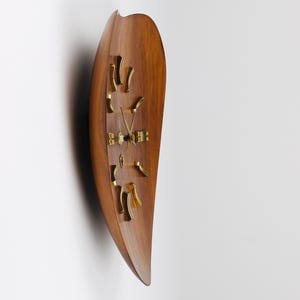 Può includere: Un orologio da parete in legno dal design curvo unico. Il quadrante dell'orologio è di colore marrone caldo, con lancette e indicatori delle ore dorati. La forma dell'orologio ricorda una foglia o una goccia, aggiungendo un tocco di eleganza moderna. Misura circa 60 cm di lunghezza.