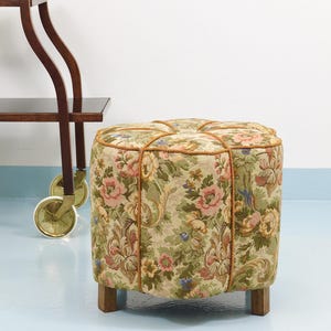 May include: A vintage floral upholstered ottoman with a square shape. The fabric features a pattern of pink, green, and yellow flowers. The ottoman is trimmed with a gold rope and has four wooden legs. A dark wood serving cart is partially visible in the background.