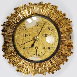 May include: A gold-toned barometer with a circular face and a cityscape design around the edge. The face has a black needle, a white dial with black markings, and the words "TIEF" and "HOCH" printed in black.