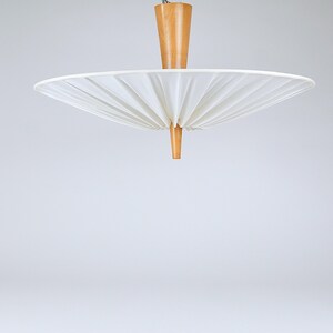 May include: A mid-century modern pendant light with a white, pleated shade and a natural wood fixture. The shade is a wide, circular shape, and the wood components have a warm, light brown color. The light fixture is suspended from the ceiling.