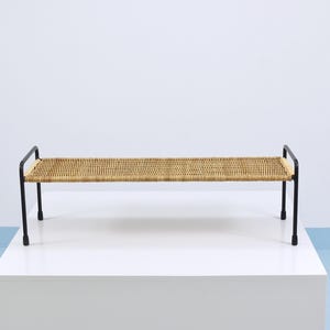 Puede incluir: Banco con asiento tejido y estructura de metal negro. El asiento es de material tejido marrón claro. El banco está sobre una superficie blanca contra una pared blanca. Mide aproximadamente 120 cm de largo.