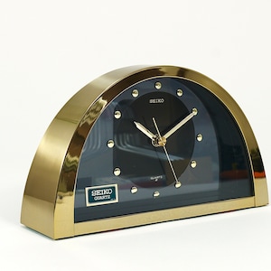 May include: A gold-coloured analogue clock with a black face and white hands. The clock is shaped like a half-circle and has the word "SEIKO" printed on the face. The clock is on a white surface.