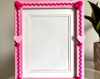 May include: Pink picture frame with a scalloped edge and pink pom-poms on the corners. The frame has a white mount and is ready for a photo.