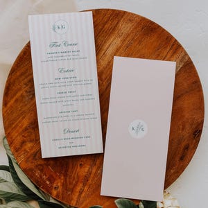 Może przedstawiać: Różowo-biała karta menu z niebiesko-zielonym wzorem kwiatowym i tekstem "KG" u góry. Menu zawiera następujące dania: Przystawka, Danie główne, Deser. W menu znajdują się: Sałatka z lokalnego rynku, Stek z Nowego Jorku, Pstrąg smażony, Tarta brioche, Tort weselny waniliowy i Asortyment makaroników.