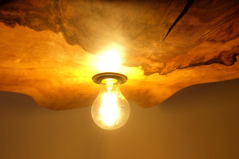 May include: A warm, glowing light bulb hangs from a wooden ceiling fixture. The wood has a natural, organic shape and a rich, golden-brown color. The light bulb is clear and emits a bright, yellow light, creating a cozy ambiance.
