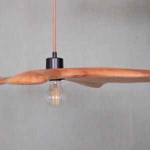 May include: A wooden pendant light fixture with a unique, organic shape. The light has a natural wood finish and a black metal fixture. The light bulb is visible and has a warm glow. The fixture hangs from a brown cord.