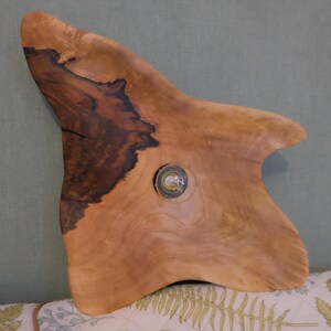 May include: A handcrafted wooden serving board with a unique, organic shape. The board features a natural wood grain pattern with dark brown and light brown tones. A small, circular, glass-covered element is embedded in the wood.