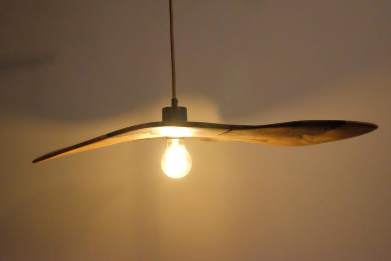 May include: A wooden, propeller-shaped pendant light fixture. The light fixture has a warm, natural wood finish and a single exposed light bulb. The fixture hangs from a thin cord, casting a warm glow.
