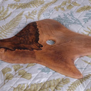 May include: A handcrafted wooden serving board with a natural, organic shape. The board features a dark, rich brown section and a lighter, reddish-brown area, with a circular hole. The board is displayed on a floral patterned fabric.