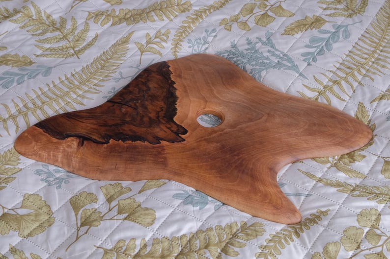 May include: A handcrafted wooden serving board with a natural, organic shape. The board features a dark, rich brown section and a lighter, reddish-brown area, with a circular hole. The board is displayed on a floral patterned fabric.