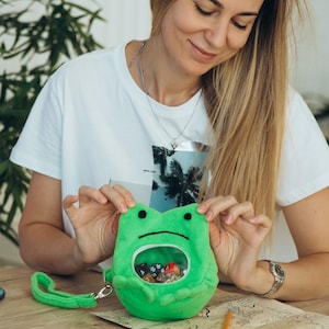 May include: A green frog-shaped dice bag, filled with dice, with a clear window and a green wrist strap. Other dice and miniature figurines are on a wooden table, along with a map and small decorative rock formations.