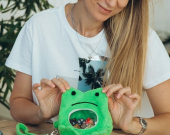 May include: A plush green frog-shaped dice bag with a clear window, filled with colorful dice. The bag has a green wrist strap. Other dice and miniature figurines are on a wooden table, along with a map and small decorative rock formations.