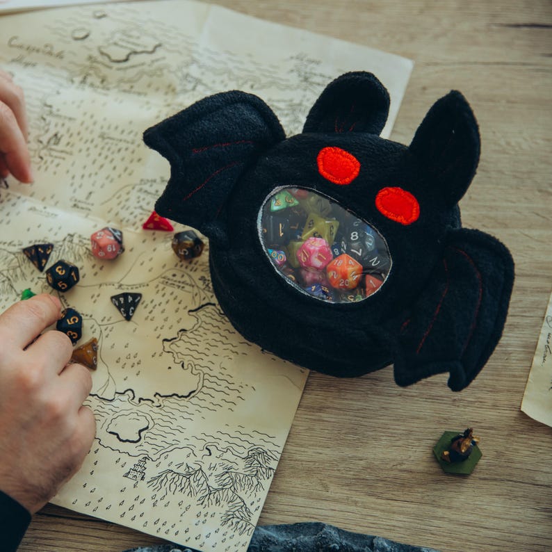 handmade dice bag price range $30 to $50 US. Handmade Dice Bag mothman crafted from soft fleece with a clear vinyl window for dice and pin display. This cryptid-themed dnd dicebags design fits 30+ dice and 3–5 pins, secured with a sturdy zipper closure. Perfect as rpg gifts for D&D fans, dungeon masters, and collectors, this mothman ita bag combines mysterious charm with practical storage. Gift-ready in eco-friendly packaging, ideal for tabletop gamers and fantasy enthusiasts.