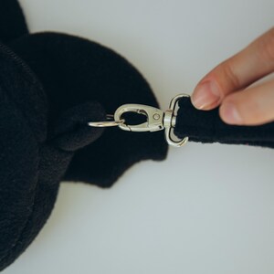 Puede incluir: Primer plano de una bolsa de felpa negra con un cierre de metal plateado y un accesorio de correa. La bolsa tiene forma de ala. Una mano est&aacute; sujetando la correa a la bolsa.