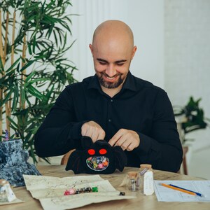 Puede incluir: Una bolsa de dados negra con forma de murci&eacute;lago con ojos rojos, llena de dados de colores. La bolsa est&aacute; abierta, revelando los dados en su interior. Una persona sonr&iacute;e y sostiene la bolsa. Un mapa y otros accesorios de juego est&aacute;n sobre la mesa.