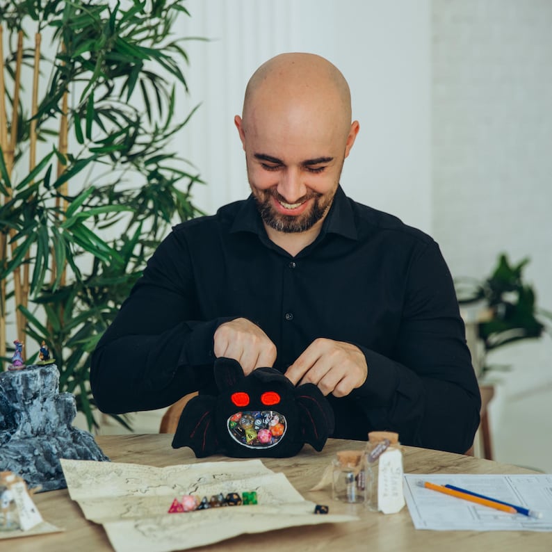 Puede incluir: Una bolsa de dados negra con forma de murci&eacute;lago con ojos rojos, llena de dados de colores. La bolsa est&aacute; abierta, revelando los dados en su interior. Una persona sonr&iacute;e y sostiene la bolsa. Un mapa y otros accesorios de juego est&aacute;n sobre la mesa.