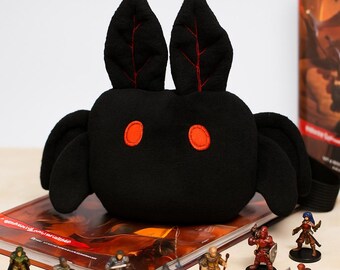 May include: A black plush toy shaped like a moth with large leaf-like ears and orange eyes. The moth is sitting on a book with miniature figurines around it. The moth's ears have red vein detailing.