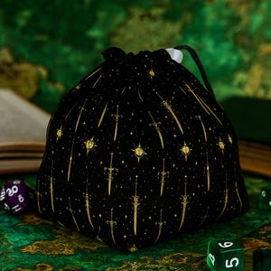 May include: Black dice bag featuring a gold sword and star design. The bag is made from a soft fabric and is intended for storing dice used in tabletop games. It has a drawstring closure and a white bead.